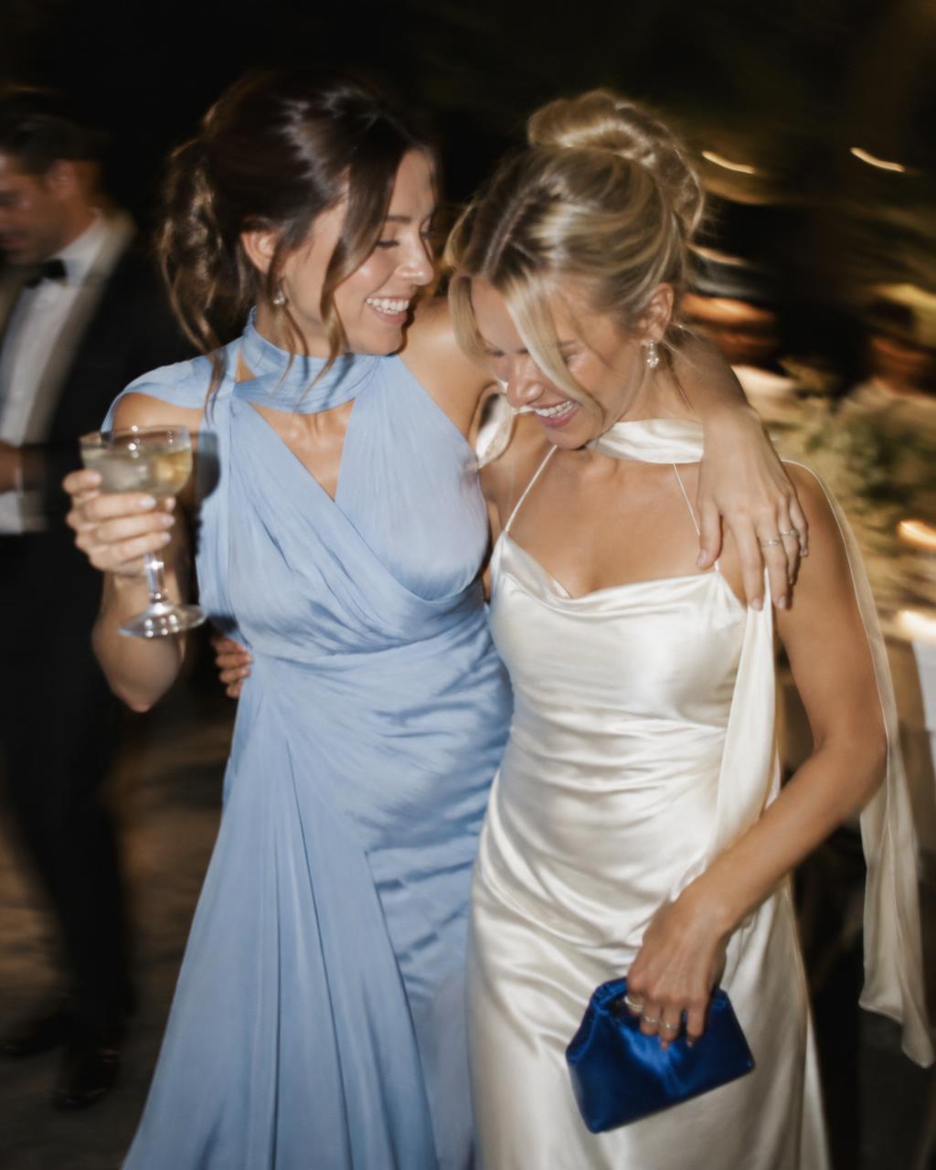 Two wedding guests walking arm in arm and laughing at an evening reception, one in a powder blue gown and one in ivory silk carrying a cobalt clutch